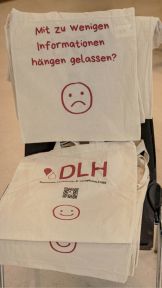 DLH-Leinentaschen mit Spruch "Mit zu wenigen Informationen hängen gelassen?" und traurigem Emoji, auf der anderen Seite DLH-Logo mit Smiley